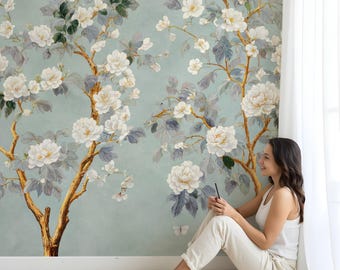 Puede incluir: Una mujer se sienta en una alfombra blanca frente a un mural de pared con un gran diseño floral con flores blancas y ramas doradas sobre un fondo azul claro.