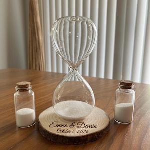Puede incluir: Un reloj de arena de cristal transparente con arena blanca, sobre una rodaja de madera grabada con "Emma & Darrin October 5, 2026." Dos pequeñas botellas de vidrio con tapones de corcho y arena blanca. Un juego de ceremonia de arena de boda.