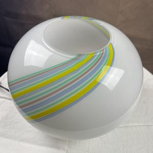 Può includere: Lampada a globo in vetro bianco con un design a strisce colorate in giallo, blu e verde. La lampada ha una forma rotonda con un'apertura nella parte superiore. Le strisce si incurvano attorno al globo.