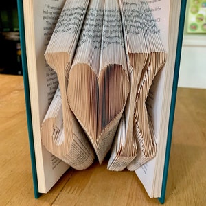 May include: A teal-covered book with pages folded to spell "J♡K". The heart shape is in the center, with the letters on either side. The book is open, revealing the folded pages and text.