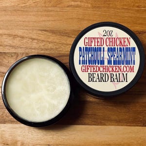 Puede incluir: Un recipiente con borde negro de bálsamo para barba junto a su tapa. La tapa muestra el texto "2oz GIFTED CHICKEN PATCHOULI SPEARMINT GIFTEDCHICKEN.COM BEARD BALM" en rojo, blanco y azul. El bálsamo es de color amarillo claro.