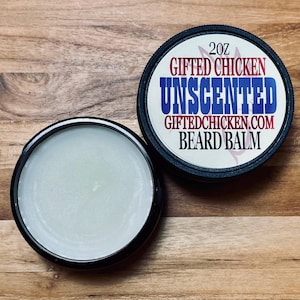 May include: Two black-rimmed containers of beard balm sit on a wooden surface. One container is open, revealing a light-colored balm. The lid of the other container displays the text "Gifted Chicken Unscented Beard Balm" and "2oz".