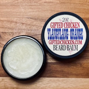 May include: Two black-rimmed containers of beard balm sit on a wooden surface. One container is open, revealing a light-colored balm. The other container's lid displays the text "Gifted Chicken Ylang Ylang Orange Beard Balm" and the website address.