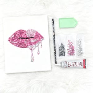 May include: A DIY diamond painting kit featuring a canvas with a pink and crystal-studded lip design. Includes rhinestones, a tray, a stylus, and a tube of B-7000 glue. The kit is designed for creating a sparkling artwork.