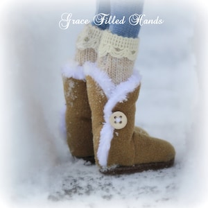 May include: A pair of brown suede boots with white faux fur trim and a single button on the side. The boots are worn by a doll with white knit socks and a blue denim skirt.