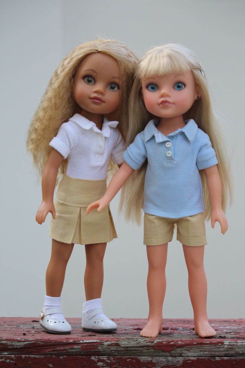 May include: Two dolls wearing school uniforms. One doll has blonde hair and is wearing a light blue polo shirt and khaki shorts. The other doll has brown hair and is wearing a white polo shirt and a khaki skirt. Both dolls are wearing white socks and shoes.