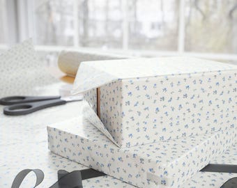 May include: Two rectangular gift boxes wrapped in white paper with a delicate blue floral pattern. One box is partially unwrapped, revealing the cardboard underneath. A pair of black scissors and a roll of wrapping paper are in the background.