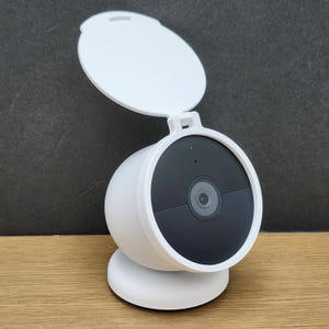 May include: A white security camera with a black lens and a protective cover. The camera is round and sits on a white base. The cover is open, revealing the lens. The camera is on a wooden surface.