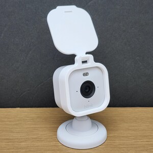May include: A white Blink security camera with a protective cover. The camera has a square body with a black lens and the brand name "blink" printed on the front. It is mounted on a round base.