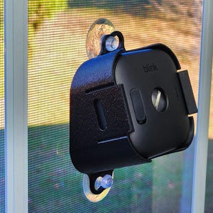 May include: A black security camera with the brand name "blink" printed on it, mounted on a window screen with suction cups. The camera is encased in a textured black housing with a rectangular shape and several openings.