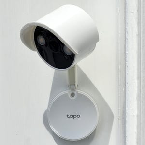 Op de afbeelding: Een witte beveiligingscamera met een beschermkap is op een witte muur gemonteerd. De camera heeft een zwarte lens en twee lampjes. Een witte, cirkelvormige basis met het woord "tapo" is eronder bevestigd.