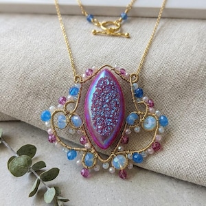 Può includere: Una collana in filo metallico dorato con una pietra druzy magenta iridescente a forma di marquise al centro. Il ciondolo è decorato con piccole perline blu, rosa e opalescenti, sospeso a una sottile catenina dorata con chiusura a T.