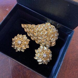 May include: A set of gold tone jewelry featuring a large leaf-shaped brooch and two matching earrings. The jewelry is adorned with clear rhinestones.