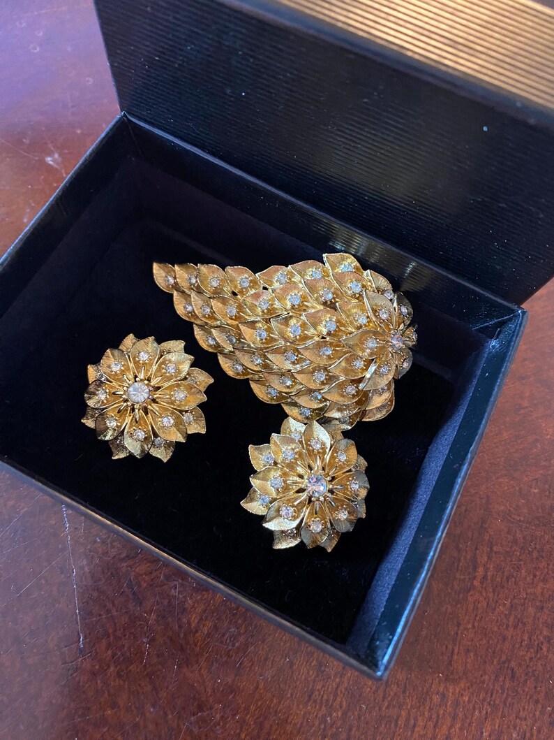 May include: A set of gold tone jewelry featuring a large leaf-shaped brooch and two matching earrings. The jewelry is adorned with clear rhinestones.