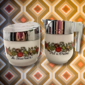 May include: A vintage cream-colored sugar bowl and creamer set. The sugar bowl has a silver lid and the creamer has a silver spout and handle. Both pieces are decorated with a colorful vegetable and fruit garland and the words "Le Sucrier" and "Pot a Creme".