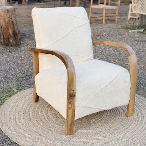 Puede incluir: Un sillón con tapicería de tela texturizada en color crema con un sutil patrón de costuras diagonales. La silla tiene reposabrazos y patas de madera curvada con un acabado de veta natural, proporcionando una estructura sólida para el asiento.
