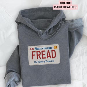 May include: Dark heather gray hoodie with a license plate graphic that reads "FREAD" in red letters. The license plate design includes the text "Massachusetts" and "The Spirit of America". The hoodie has a hood and long sleeves.