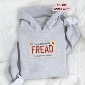 May include: A light gray hoodie with a hood and long sleeves. The front of the hoodie features a patch resembling a Massachusetts license plate with the name "FREAD" in red letters. The text "The Spirit of America" is below the name.