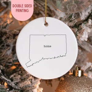 Peut inclure: Une boule de Noël en céramique blanche avec un contour noir de l'État du Connecticut et le mot "home". L'ornement a une ficelle dorée pour l'accrocher et est présenté sur un sapin de Noël. Le texte "DOUBLE SIDED PRINTING" est dans un ovale rose.
