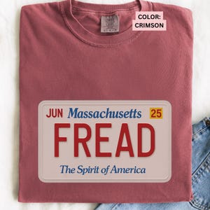 May include: Crimson-colored t-shirt with a Massachusetts license plate design. The license plate reads "FREAD" in red letters, with "The Spirit of America" below. The shirt is a Comfort Colors brand.