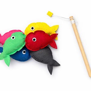 May include: A colorful felt fishing game set with six fish in various colors: pink, green, blue, yellow, red, and gray. A wooden fishing rod with a string and a yellow hook is included. The fish have white eyes.
