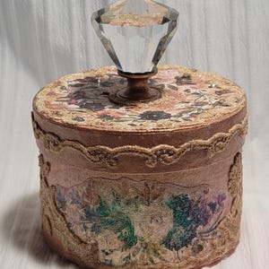 May include: A decorative round box with a clear, faceted crystal knob. The box is adorned with floral patterns and gold trim. The sides feature a floral design, and the box has a pinkish-brown hue.