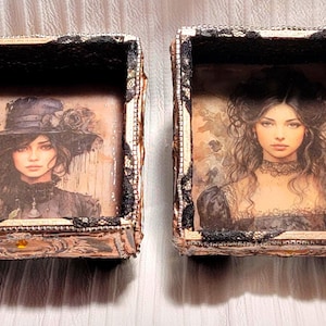 May include: Two decorative boxes with vintage-style portraits of women. The boxes have a metallic finish, black lace trim, and ornate details. One portrait features a woman in a hat, the other a woman with curly hair.