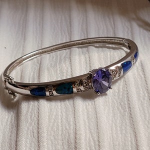 May include: A silver bangle bracelet with a purple gemstone and blue and green opal accents.