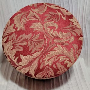 May include: A round, red fabric ottoman with a beige floral pattern. The ottoman is covered in a textured fabric.