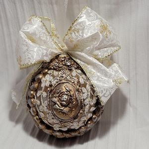 May include: Ornate Christmas ornament with a gold and bronze finish, featuring a floral design and a cameo with a rose. A white bow with gold trim and a floral pattern adorns the top, adding a touch of elegance to this holiday decoration.