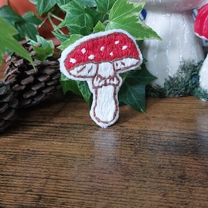 Könnte beinhalten: Ein weiß gestickter Pilz mit roter Kappe und weißen Punkten. Der Pilz befindet sich auf einer braunen Holzoberfläche.