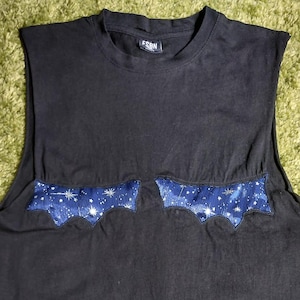 Può includere: Una canotta nera con scollo rotondo e due applicazioni in tessuto a forma di pipistrello sul petto. Le applicazioni sono realizzate in tessuto blu scuro con un motivo di stelle e costellazioni bianche. Una piccola etichetta nera è visibile sul collo.