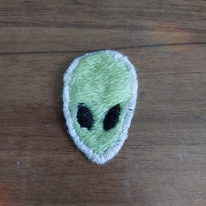 Könnte beinhalten: Ein gestickter Aufnäher in Form eines Alien-Kopfes. Der Aufnäher zeigt ein hellgrünes Gesicht mit zwei großen, ovalen schwarzen Augen. Das gesamte Design ist mit einem dicken, weißen gestickten Rand umrandet, was einen deutlichen Kontrast erzeugt.
