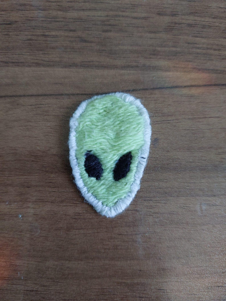 Peut inclure: Un &eacute;cusson brod&eacute; en forme de t&ecirc;te d'extraterrestre. L'&eacute;cusson pr&eacute;sente un visage vert clair avec deux grands yeux noirs de forme ovale. L'ensemble du motif est soulign&eacute; par une bordure &eacute;paisse cousue en blanc, cr&eacute;ant un contraste marqu&eacute;.