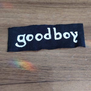 Könnte beinhalten: Ein rechteckiger schwarzer Stoffaufnäher mit dem in weißem Garn gestickten Wort goodboy. Die Schrift ist in einem kleingeschriebenen, abgerundeten Stil gehalten. Der Aufnäher hat raue, leicht unebene Kanten und liegt horizontal.