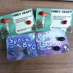 Op de afbeelding: Verschillende handgemaakte patches en spelden worden getoond. De spelden tonen lieveheersbeestjes en ruimte-thema insecten. De kaarten lezen "Piney Craft" en "Space Bugs From Space!" met social media informatie.