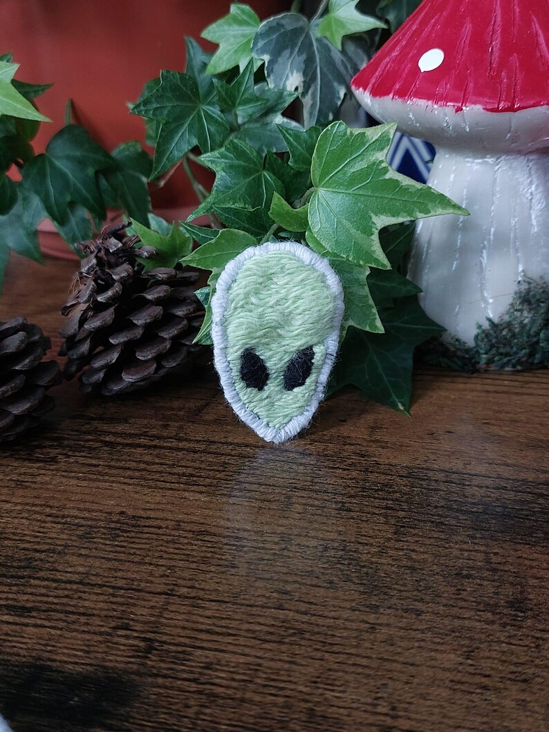 K&ouml;nnte beinhalten: Ein hellgr&uuml;ner Alien-Aufn&auml;her mit schwarzen Augen und wei&szlig;em Rand. Der Aufn&auml;her befindet sich vor gr&uuml;nem Efeu und einem dekorativen Pilz mit rotem Hut. Zwei Tannenzapfen sind ebenfalls zu sehen.