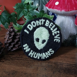 Könnte beinhalten: Ein runder, schwarzer Aufnäher mit dem weißen Text "I DON'T BELIEVE IN HUMANS". Der Aufnäher zeigt einen grünen Alienkopf in der Mitte. Dekorative Pilze und Blätter sind im Hintergrund.