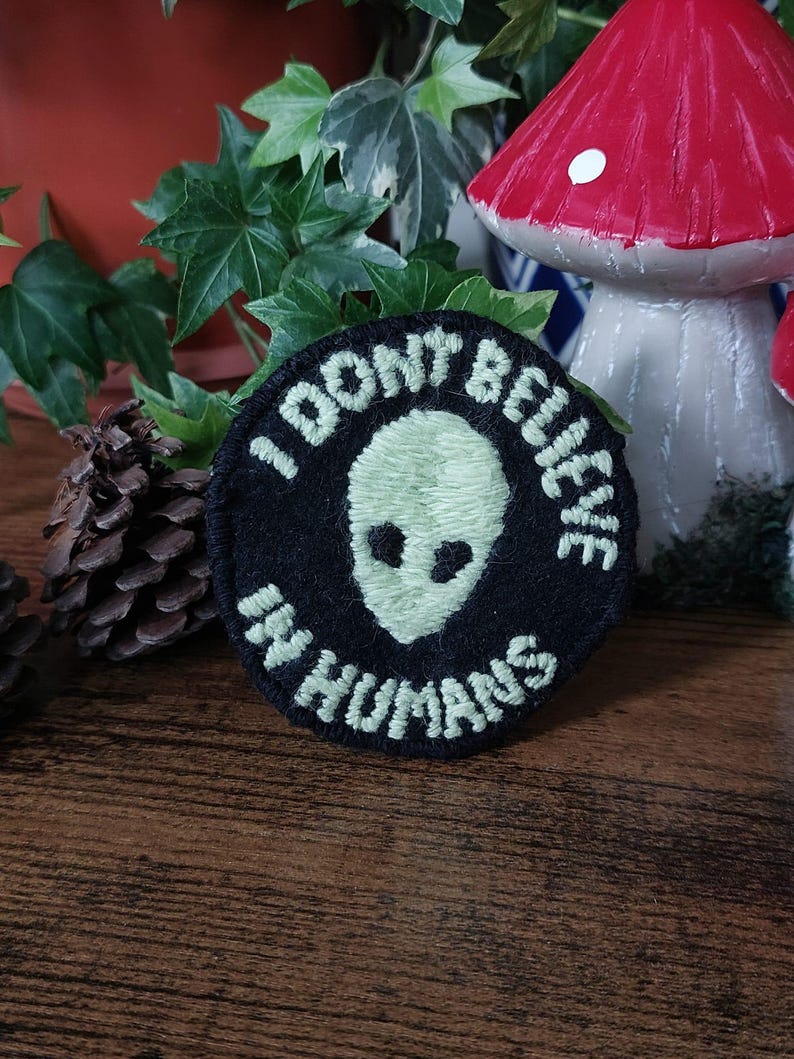 K&ouml;nnte beinhalten: Ein runder, schwarzer Aufn&auml;her mit dem wei&szlig;en Text "I DON'T BELIEVE IN HUMANS". Der Aufn&auml;her zeigt einen gr&uuml;nen Alienkopf in der Mitte. Dekorative Pilze und Bl&auml;tter sind im Hintergrund.