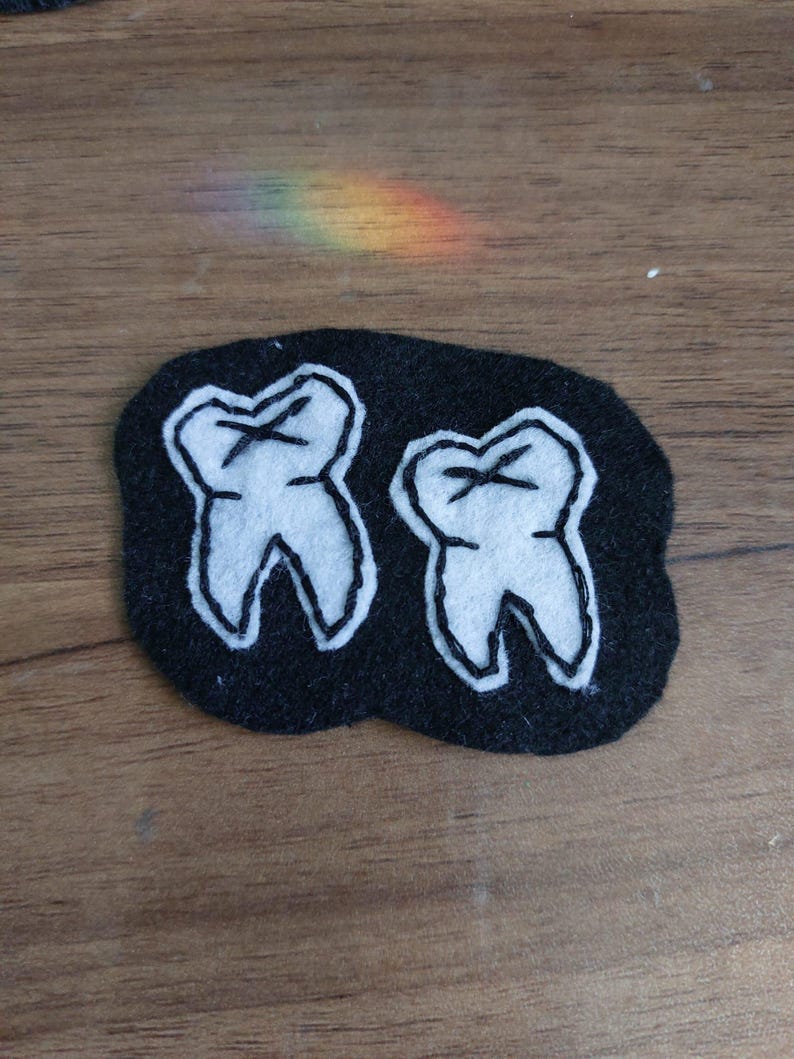 May include: A black felt patch featuring two white embroidered teeth. Each tooth is outlined in black thread with a black cross-stitch detail on the crown. The patch has an irregular, rounded shape and is crafted from soft, layered fabric materials.