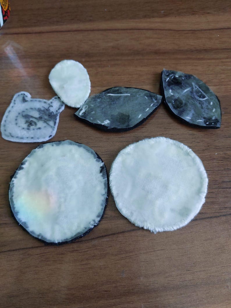 May include: A collection of six assorted fabric patches in various shapes including circles, ovals, and a character-like silhouette. The patches feature white, off-white, and dark grey colors with visible stitching along the edges and adhesive backings.