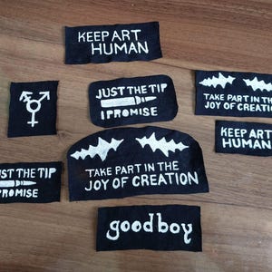 Könnte beinhalten: Eine Sammlung schwarzer Stoffaufnäher mit weiß handgemalten Motiven und Texten. Die Aufnäher zeigen Schriftzüge wie KEEP ART HUMAN, JUST THE TIP I PROMISE, TAKE PART IN THE JOY OF CREATION, good boy sowie ein Gender-Symbol.