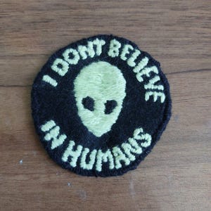 Peut inclure: Un &eacute;cusson brod&eacute; noir de forme circulaire avec une t&ecirc;te d'extraterrestre vert p&acirc;le au centre. Le texte I DONT BELIEVE IN HUMANS est brod&eacute; en fil vert p&acirc;le autour de la t&ecirc;te, en suivant le bord circulaire de l'&eacute;cusson.