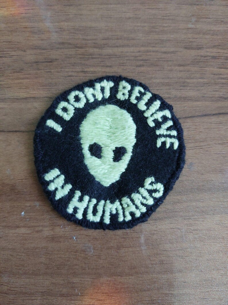 Peut inclure: Un &eacute;cusson brod&eacute; noir de forme circulaire avec une t&ecirc;te d'extraterrestre vert p&acirc;le au centre. Le texte I DONT BELIEVE IN HUMANS est brod&eacute; en fil vert p&acirc;le autour de la t&ecirc;te, en suivant le bord circulaire de l'&eacute;cusson.