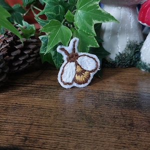 Op de afbeelding: Een witte en bruine geborduurde patch van een vuurvliegje met gele accenten. De patch bevindt zich op een houten oppervlak.