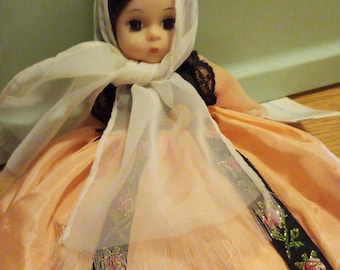 May include: A porcelain doll wearing a peach-colored dress with a white lace shawl and black trim. The doll has dark hair and brown eyes.