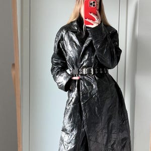 May include: A long, black, crinkled-texture coat with a wide collar and long sleeves. The coat is cinched at the waist with a black belt featuring silver-toned metal eyelets and a rectangular buckle. The coat is worn over light blue denim jeans.