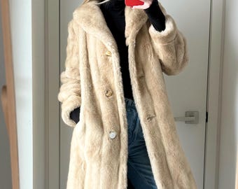 May include: A beige faux fur coat with a wide collar and three front buttons, worn over a black long-sleeve top and light blue wide-leg denim jeans. The coat features a textured, plush finish and reaches mid-thigh length.