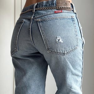 May include: Light blue denim jeans with a distressed patch on the back pocket. The jeans have a high waist and a silver chain belt. The Wrangler brand label is visible on the back.