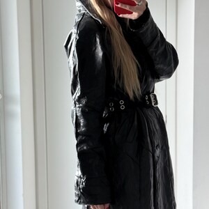 May include: Black leather trench coat with a belted waist and a high collar. The coat has a textured appearance and features a double-breasted design. The coat is a long length and appears to be vintage.